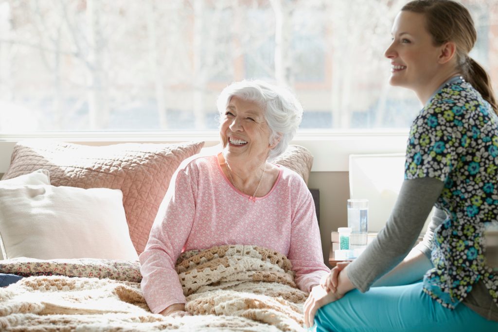 An elderly woman sits on a bed smiling at a nurse who smiles back.