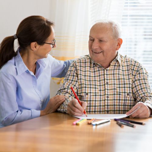 An elderly man is happily coloring next to a woman who has her arm around him and is also smiling