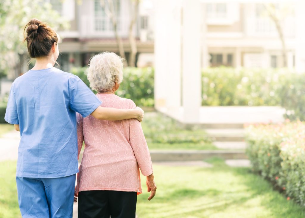 A nurse and elderly woman stand side by side outside with an arm around each other