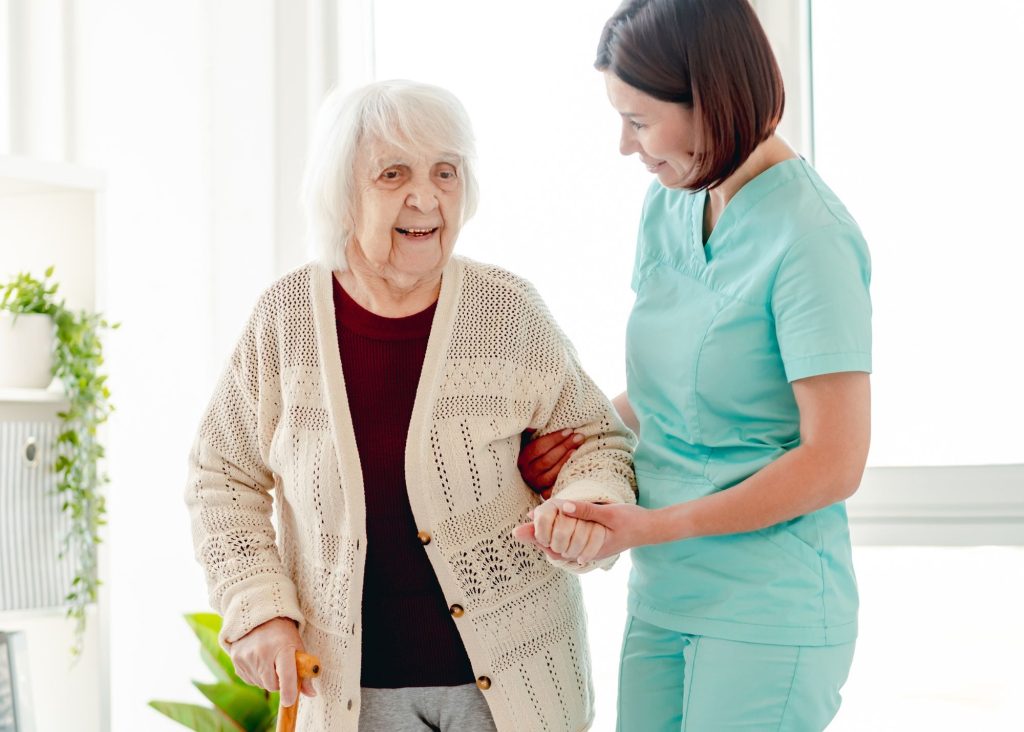 An elderly woman is walking with aid of a nurse. They are both smiling.