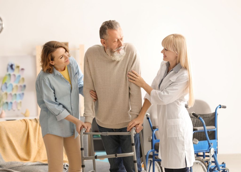 An elderly man with a walker is assisted by two smiling middle aged women.